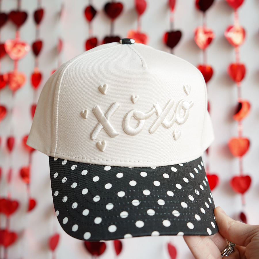 White cap with 'XOXO' text held in front of a heart-patterned background