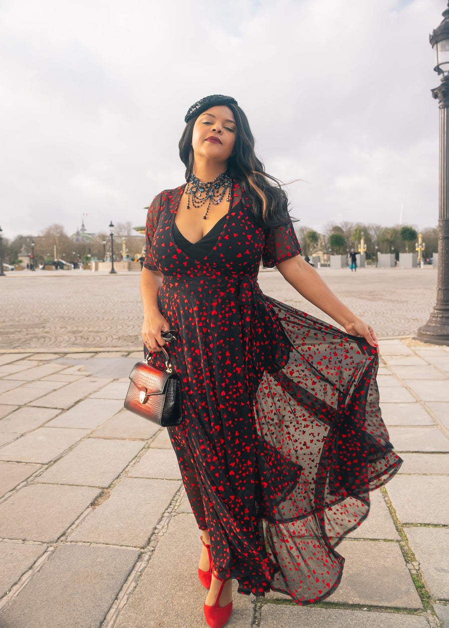 Woman in a black and red dress with sheer overlay standing on a paved area.