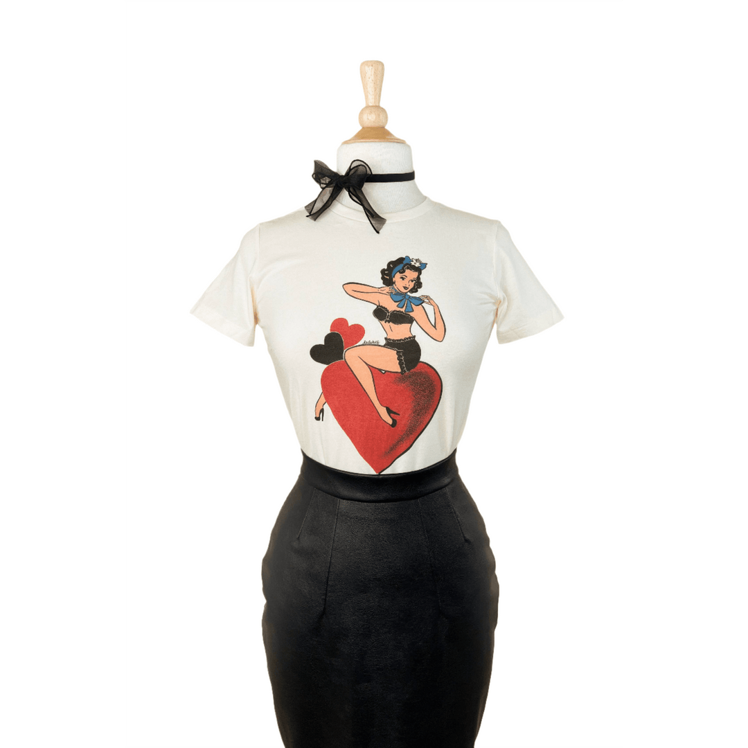 A fitted tee in ivory color with a graphic print of a character sitting on a heart, displayed on a mannequin paired with a black skirt.