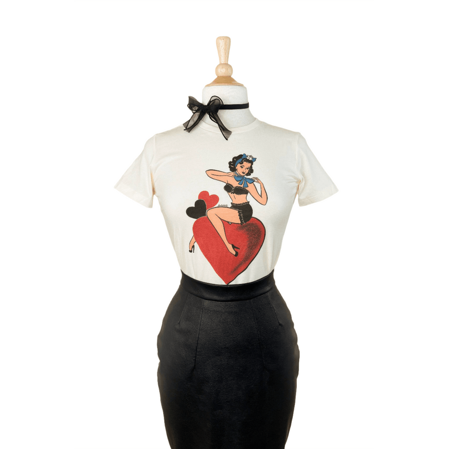 A fitted tee in ivory color with a graphic print of a character sitting on a heart, displayed on a mannequin paired with a black skirt.