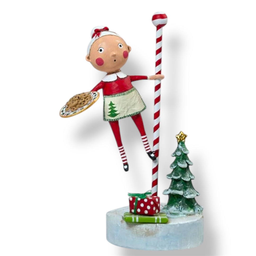 A Christmas figurine featuring a whimsical character with a red and white striped staff, a green skirt with a Christmas tree design, and holding a cookie, placed next to a miniature Christmas tree and present on a snowy base.