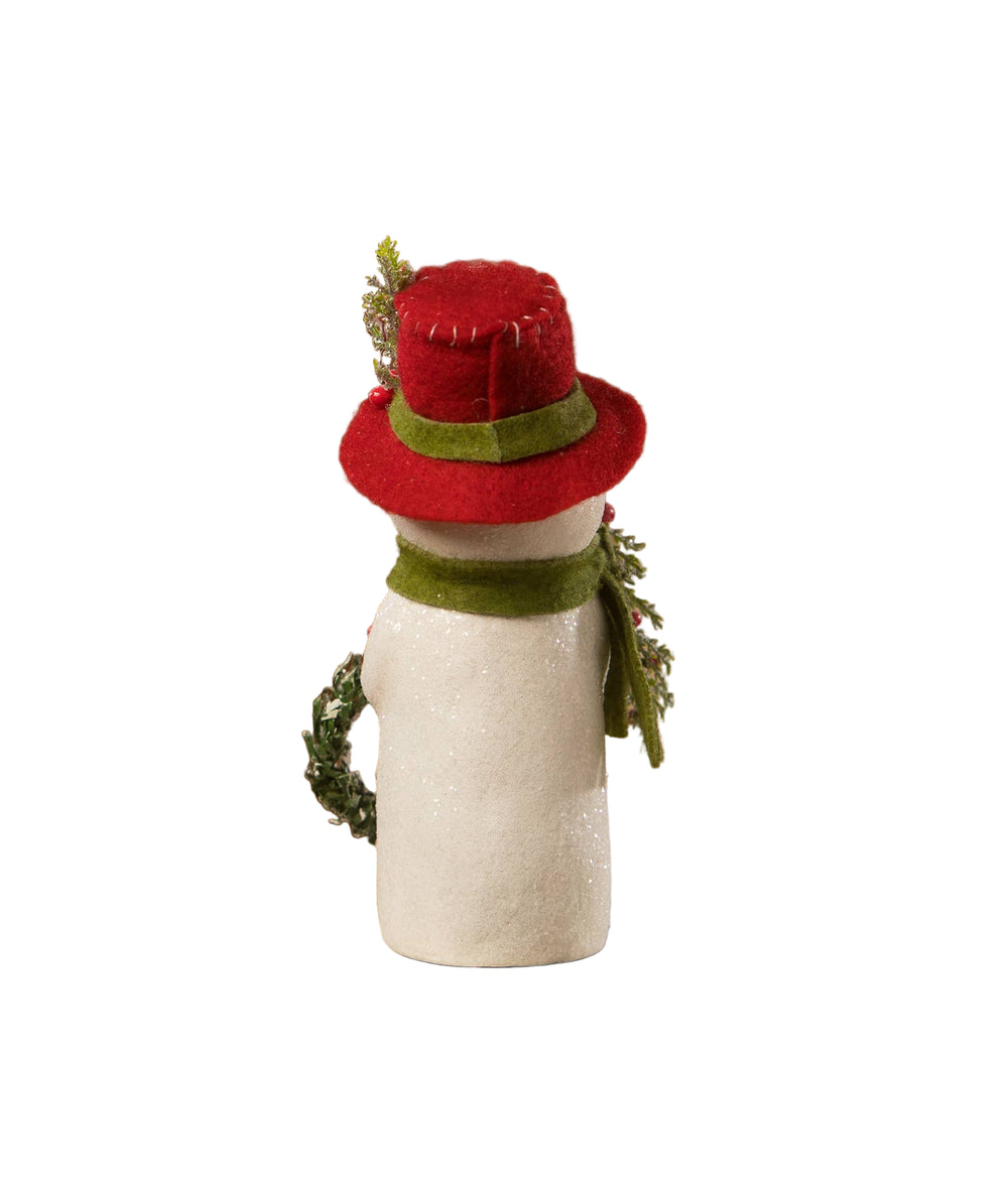 Small snowman figurine with a red hat and green scarf on a rustic wooden surface with a blurred Christmas tree in the background.
