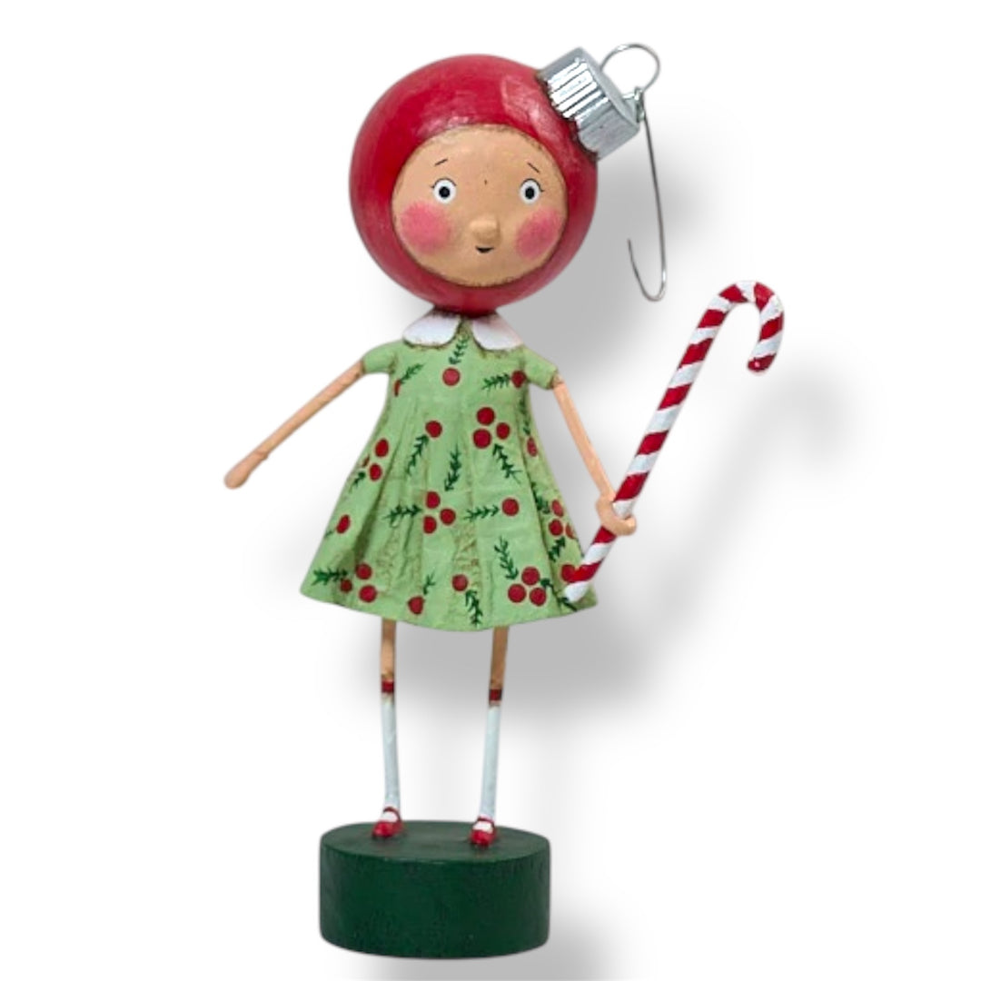 A Christmas figurine of a girl with red christmas ornament hat, wearing a green dress with a cherry pattern, holding a candy cane, and wearing a silver ornament as a hat.