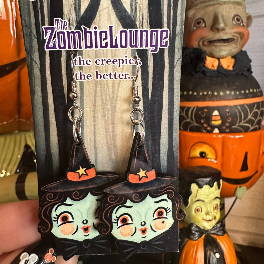 Witch-themed earrings in packaging with 'The Zombie Lounge' branding, surrounded by Halloween decorations.