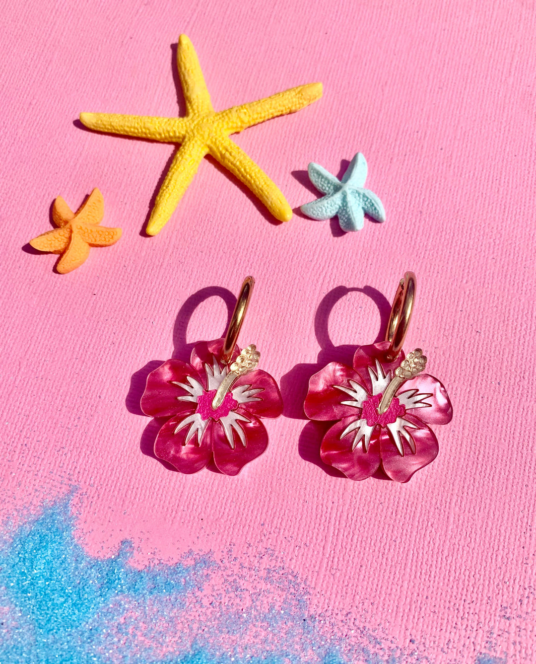 Pink flower-shaped earrings with gold hooks on a pink background with starfish decorations.