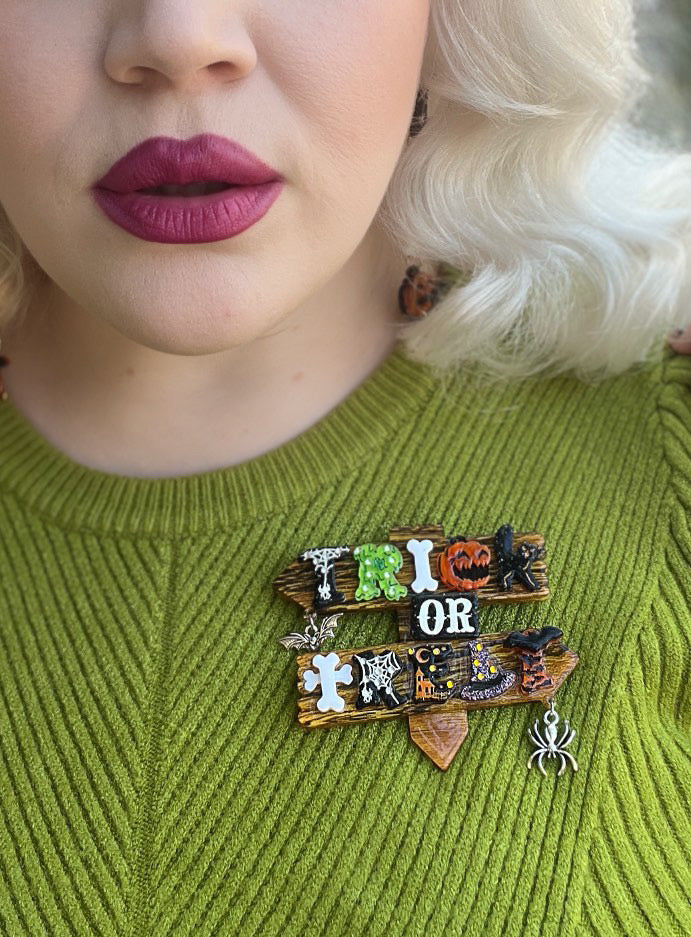 trick or Treat Brooch by Lipstick & Chrome 
Step right into spooky season with the Trick or Treat Brooch by Lipstick &amp; Chrome — a showstopping accessory designed to capture the playful magic of Halloween night. This layered acrylic brooch spells out “Trick or Treat” in bold festive letters, each decorated with pumpkins, cobwebs, bones, and bats, while dangling bat and spider charms add extra movement and flair.