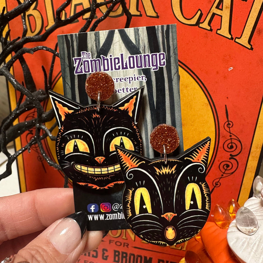 Black cat-shaped earrings with orange accents on a Zombielounge card against an orange background.