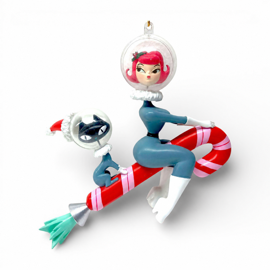 A collectible holiday ornament featuring a stylized retro illustration of a woman in a clear space helmet with pink hair, sitting on a red-and-white striped candy cane while holding a small black-and-white cat in a Santa hat, photographed on a clean white background.