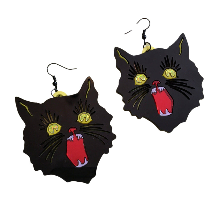 A pair of black cat-shaped statement earrings with yellow eyes and red details, displayed on a green and white fibrous surface with scattered clear crystals.