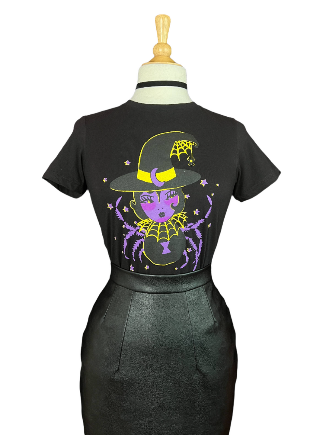 A black short-sleeve top displayed on a dress form, featuring a large back graphic of a purple witch character with a yellow hat, spiderwebs, and stars, paired with a black skirt against a white background.